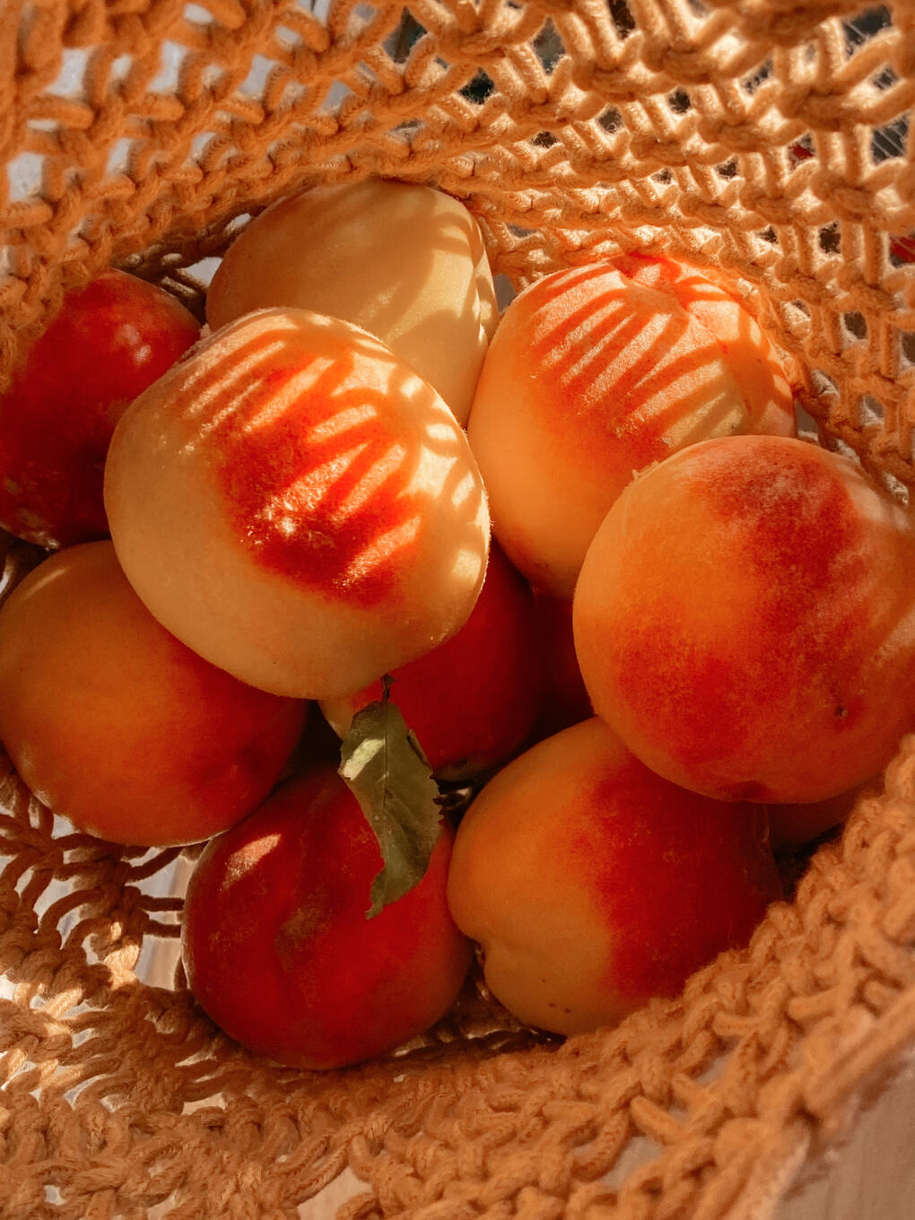 A collection of yellow and orange peaches in a woven basket with bright sunlight shining from the upper right corner